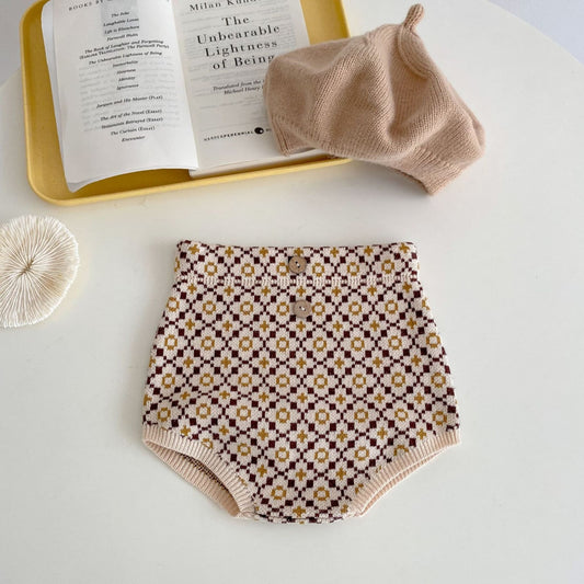 Patterned baby brown knit bloomers with a matching hat on a white table next to an open book.