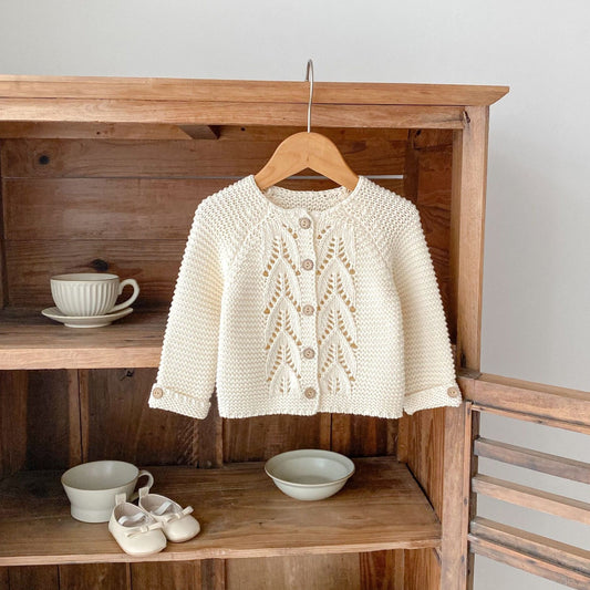 Knitted baby jacket on a hanger against a wooden background with bowls and shoes.