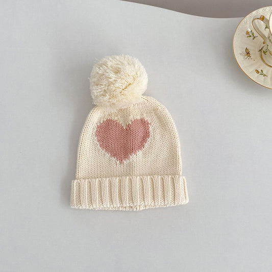 Knitted cream baby hat with a heart design and pom-pom on a light grey background