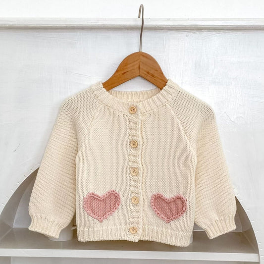 Cream knitted baby cardigan with pink heart designs on a wooden hanger against a white background