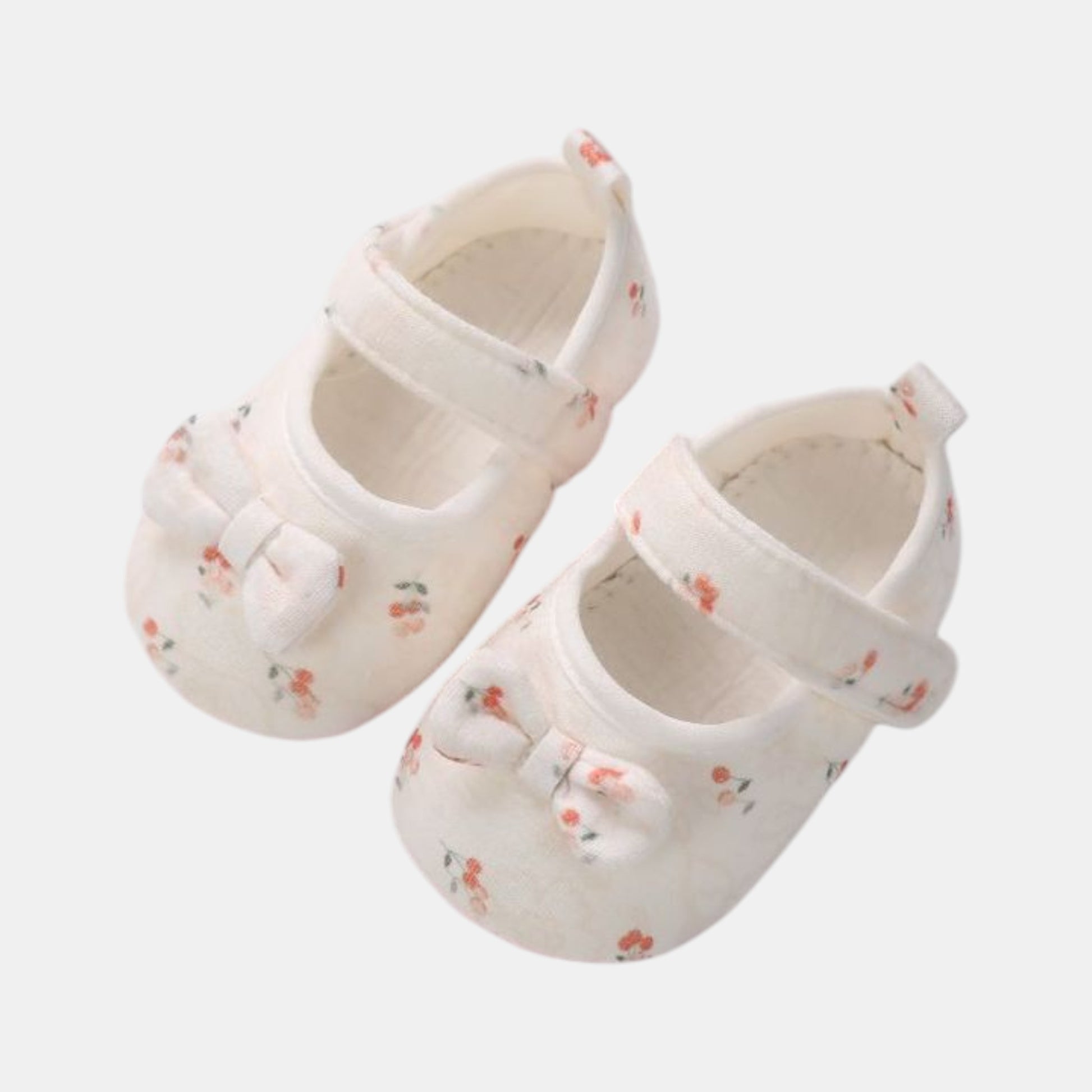 A pair of white cotton baby shoes with red cherry patterns and bows, featuring a velcro strap and grips on the sole.