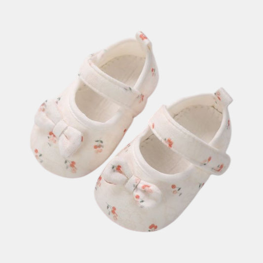 A pair of white cotton baby shoes with red cherry patterns and bows, featuring a velcro strap and grips on the sole.