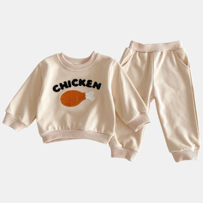 A beige two-piece lounge set for children, featuring a long-sleeve sweatshirt with 'CHICKEN' text and a drumstick illustration, and matching pants with pockets.