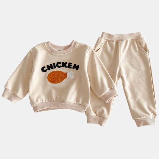 A beige two-piece lounge set for children, featuring a long-sleeve sweatshirt with 'CHICKEN' text and a drumstick illustration, and matching pants with pockets.