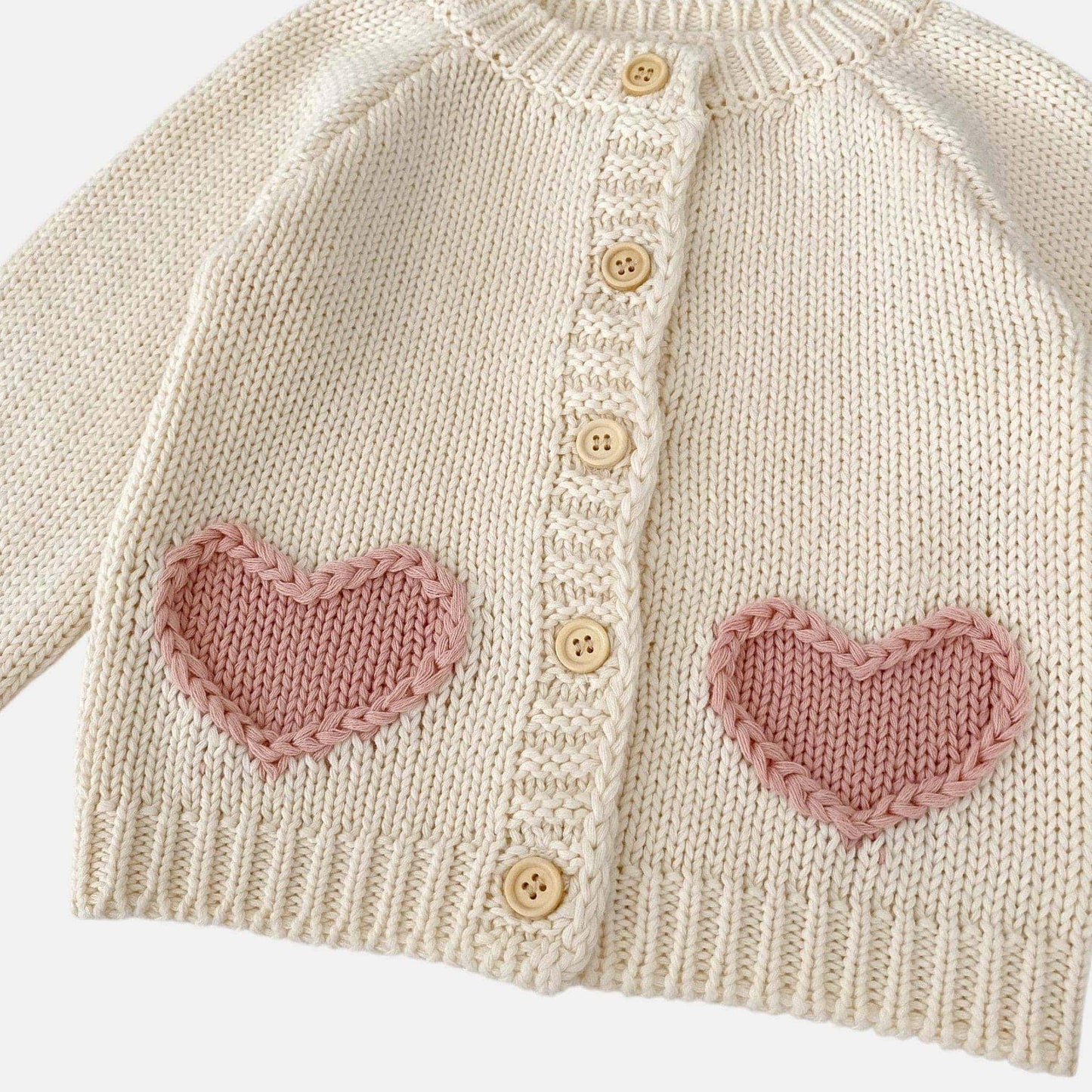 Knitted cream baby cardigan with pink heart patterns and buttons on a white background
