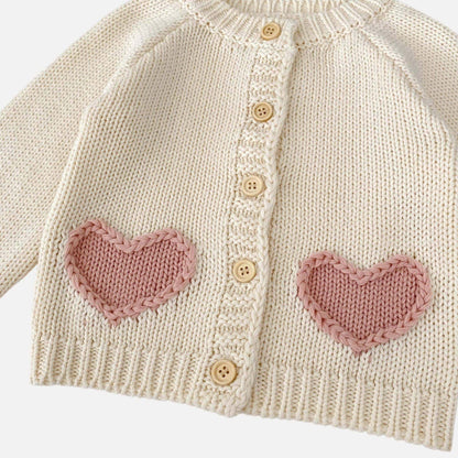 Knitted cream baby cardigan with pink heart patterns and buttons on a white background