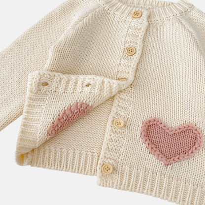 Knitted cream baby cardigan with pink heart patterns on a light background