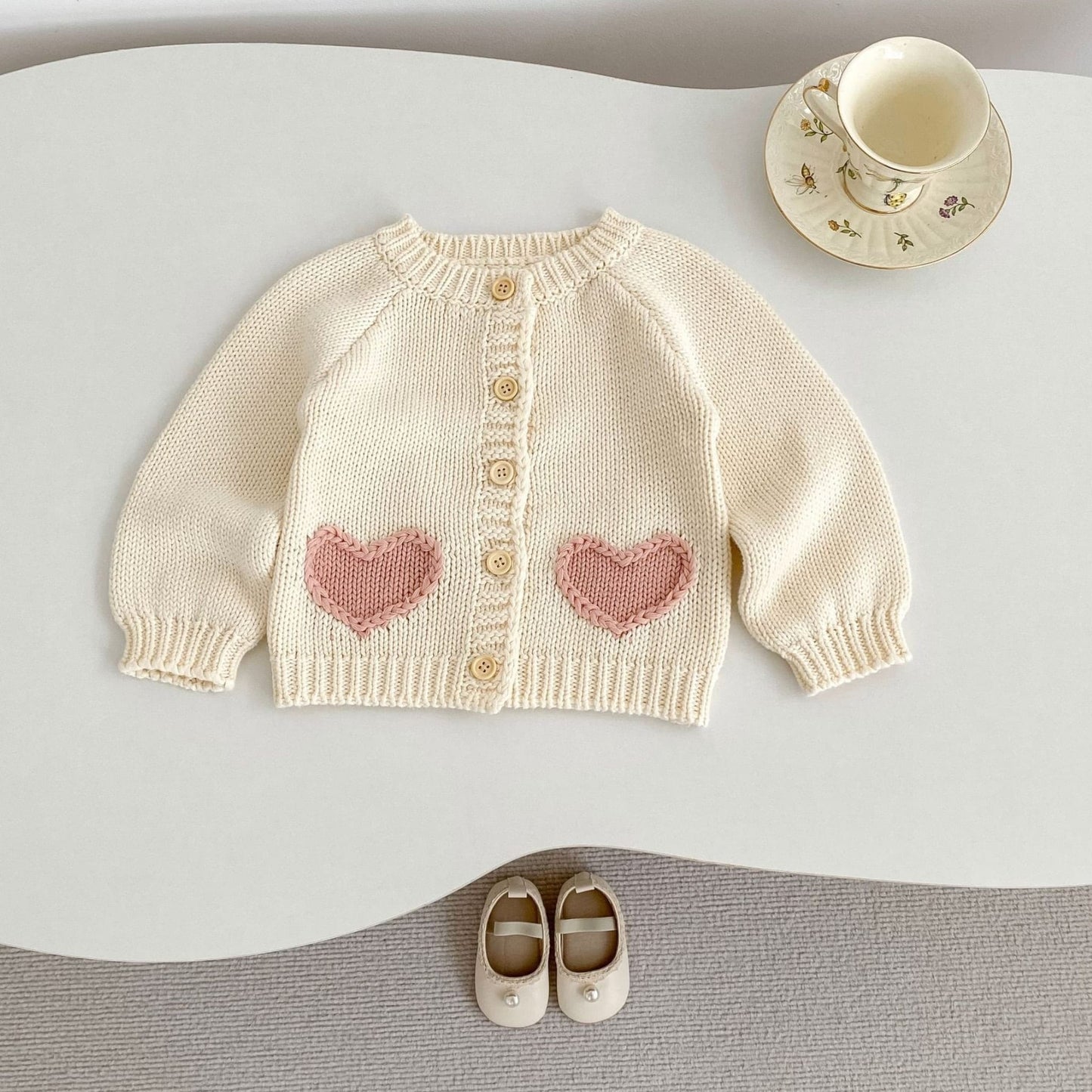 Baby cream sweater with pink heart patterns on a white surface with shoes and a cup.