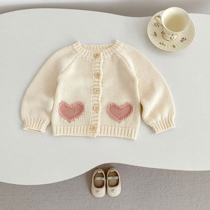Baby cream sweater with pink heart patterns on a white surface with shoes and a cup.